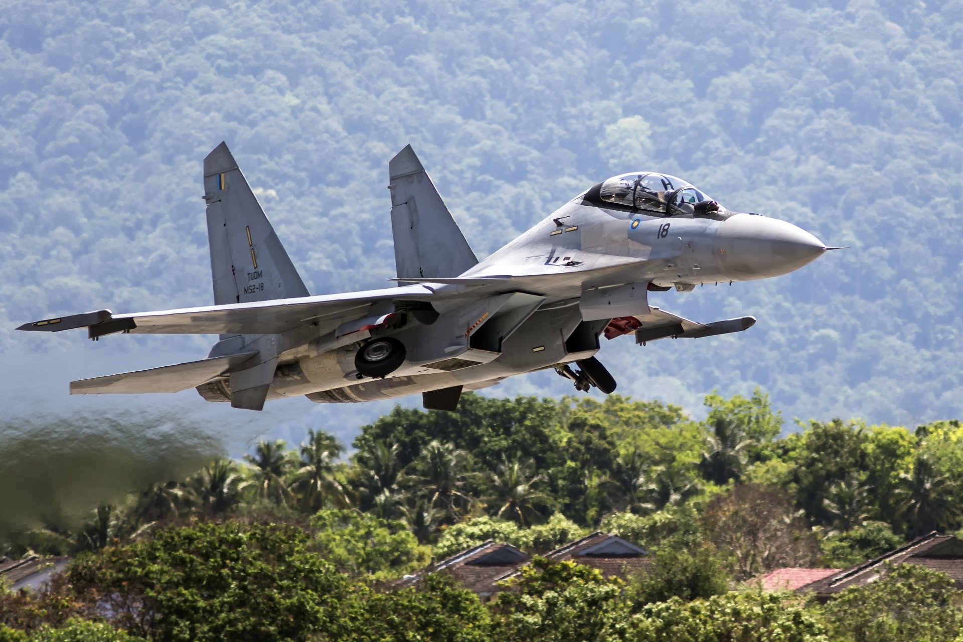 A Sukhoi Su-30 jet fighter warplane takes off over lush greenery, captured in vivid 4K Ultra HD as a military aircraft soars against a mountainous backdrop.