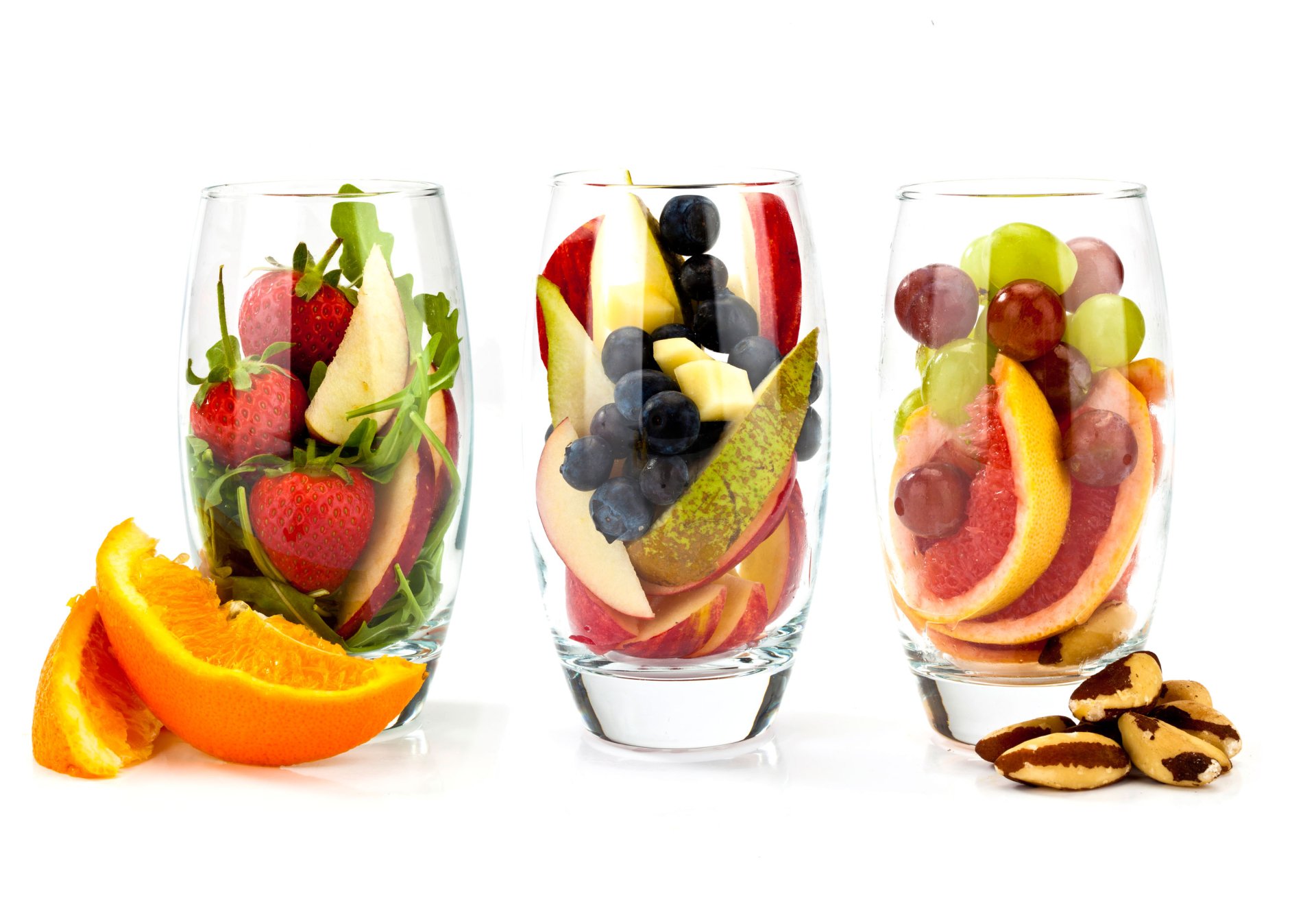 Three glasses filled with strawberries, blueberries, grapes, pear slices, and orange wedges, accompanied by orange slices and Brazil nuts on a white background.