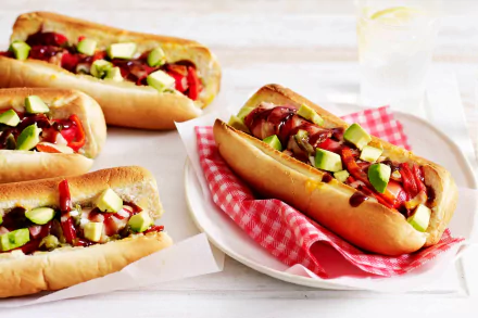 Bright 2K Quad HD PC desktop wallpaper of loaded hot dogs with avocado, chopped tomatoes and sauce on soft buns, arranged on plates with a red gingham napkin.