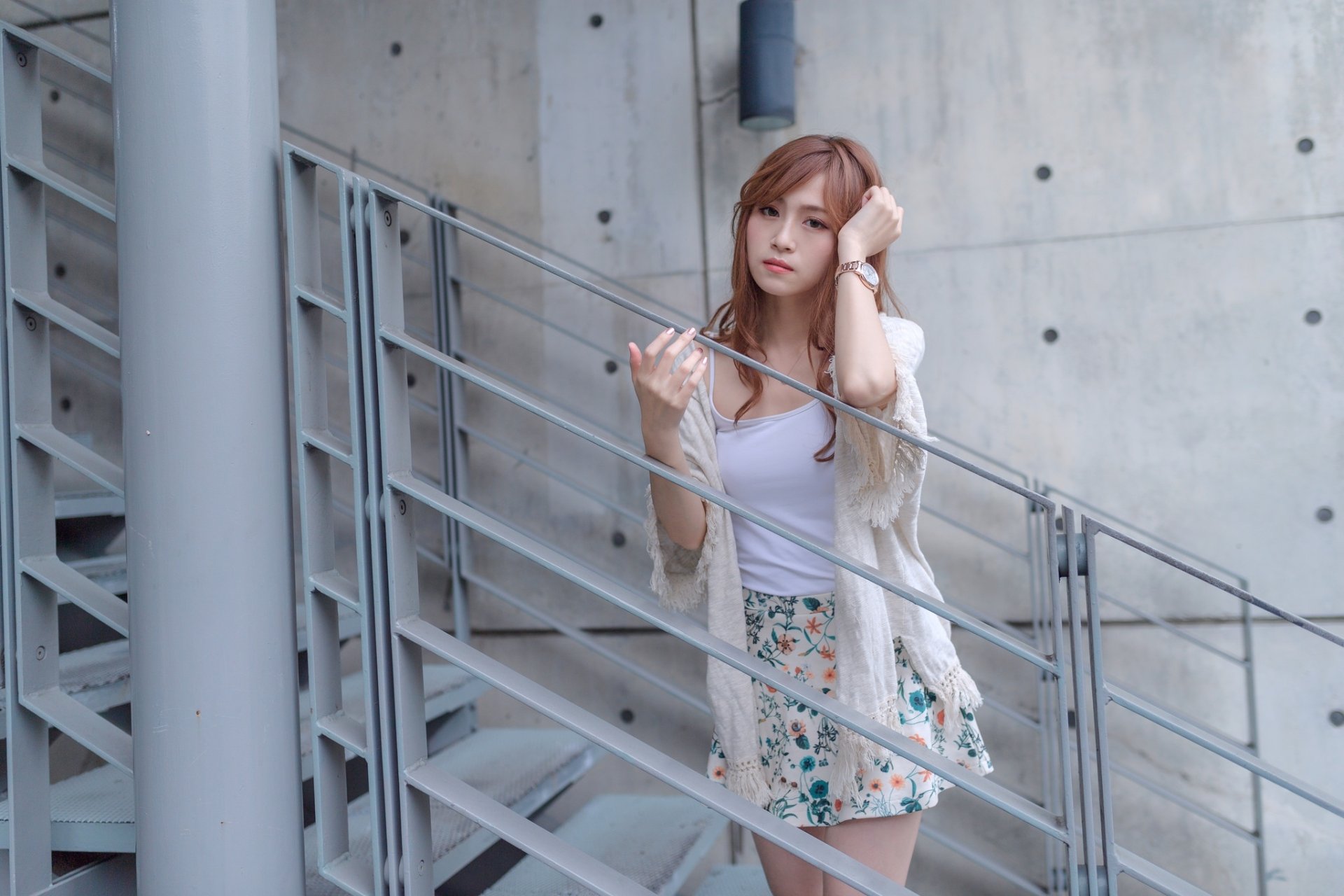 HD desktop wallpaper of an Asian redhead woman wearing a floral skirt and white top, posing thoughtfully on a metal staircase against a concrete wall background.