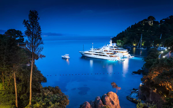 HD desktop wallpaper of a serene night view in Portofino, Italy, featuring a luxurious yacht anchored in the calm waters, surrounded by lush greenery and illuminated by soft lights.