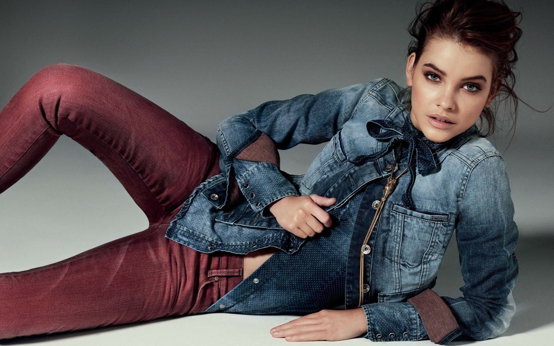 Brunette model Barbara Palvin with striking blue eyes stares confidently while reclining, dressed in a denim jacket and maroon jeans against a neutral background.