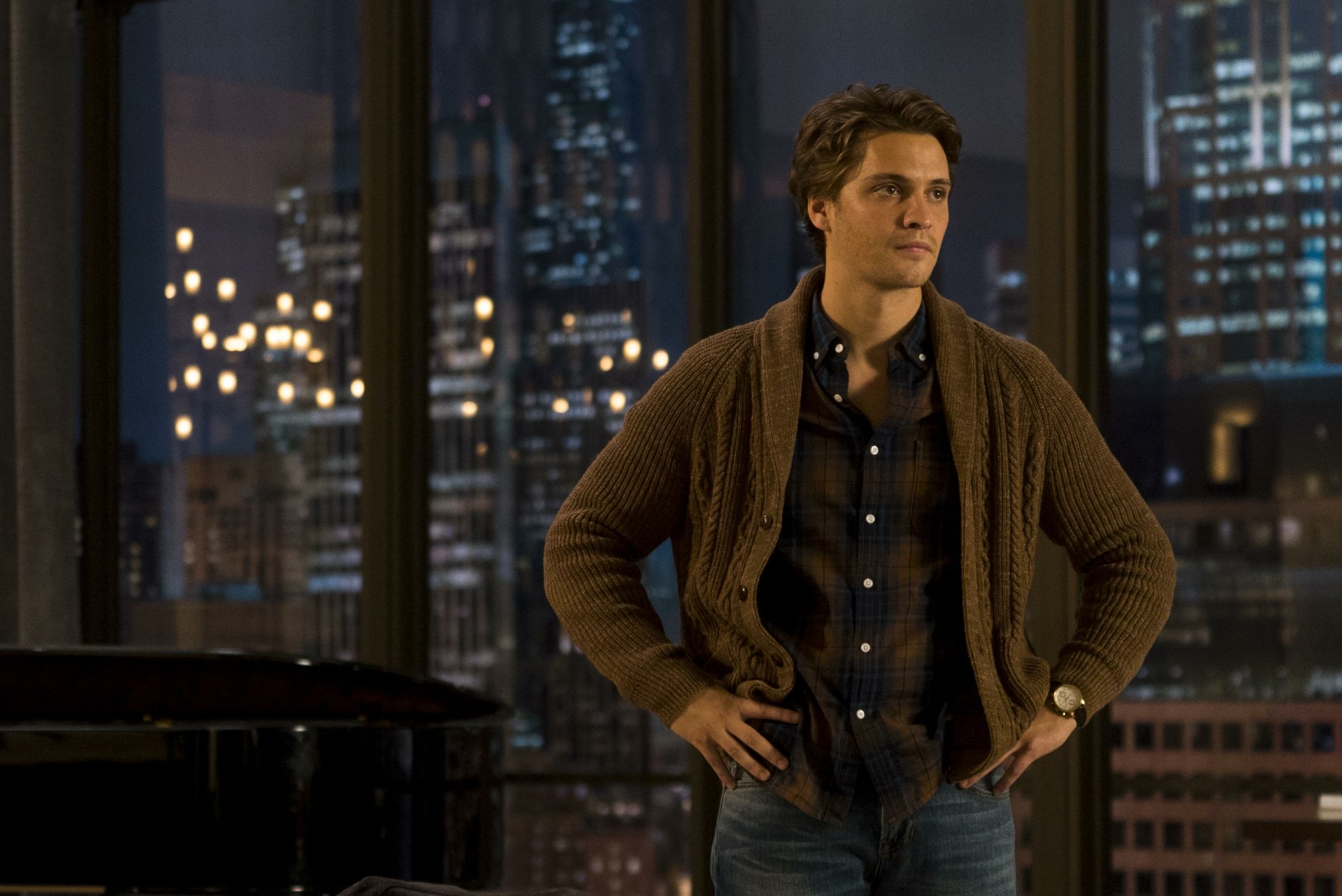 HD desktop wallpaper featuring Luke Grimes from the movie Fifty Shades Darker. He stands confidently in front of a cityscape backdrop, wearing a brown sweater and a plaid shirt.