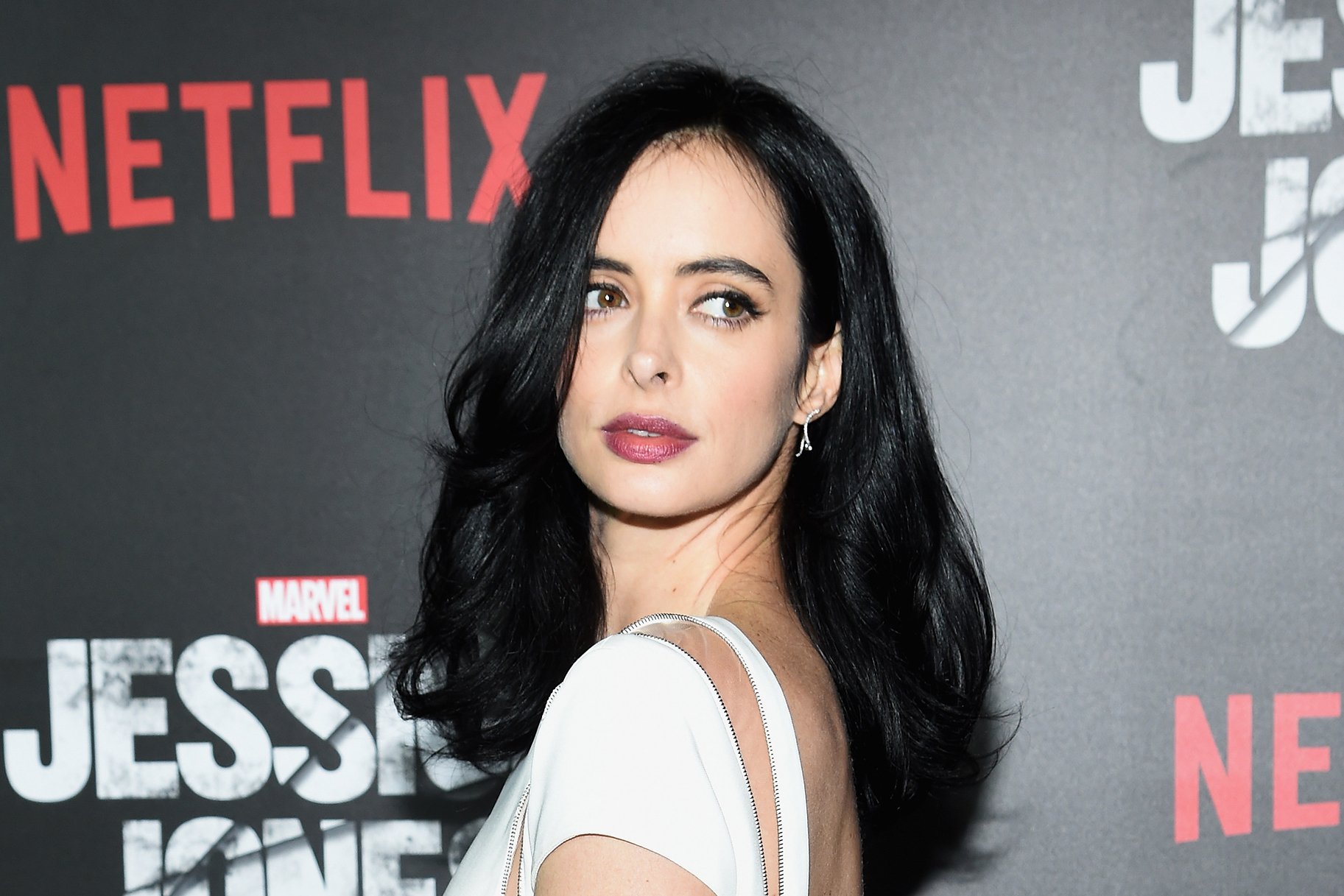 HD desktop wallpaper of a dark-haired woman in a white dress looking over her shoulder against a red-and-black premiere backdrop with Netflix and Jessica Jones logos.