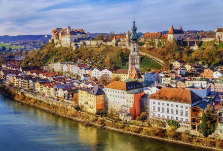 Scenic view of Burghausen, Bavaria, Germany, featuring a man-made town along the river with historic buildings and a fortress on the hill under a cloudy sky.