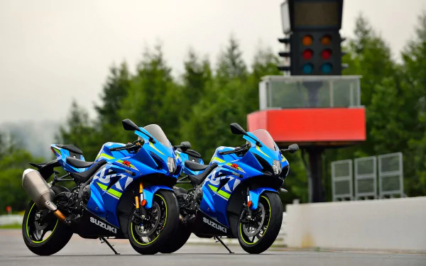 HD desktop wallpaper featuring two blue Suzuki GSX-R1000 motorcycles parked on a racetrack with a blurred background of trees and a traffic light tower.
