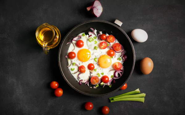 4K Ultra HD still life of a frying pan with cooked eggs, cherry tomatoes, and green onions, surrounded by whole eggs, garlic, and tomatoes on a dark surface.
