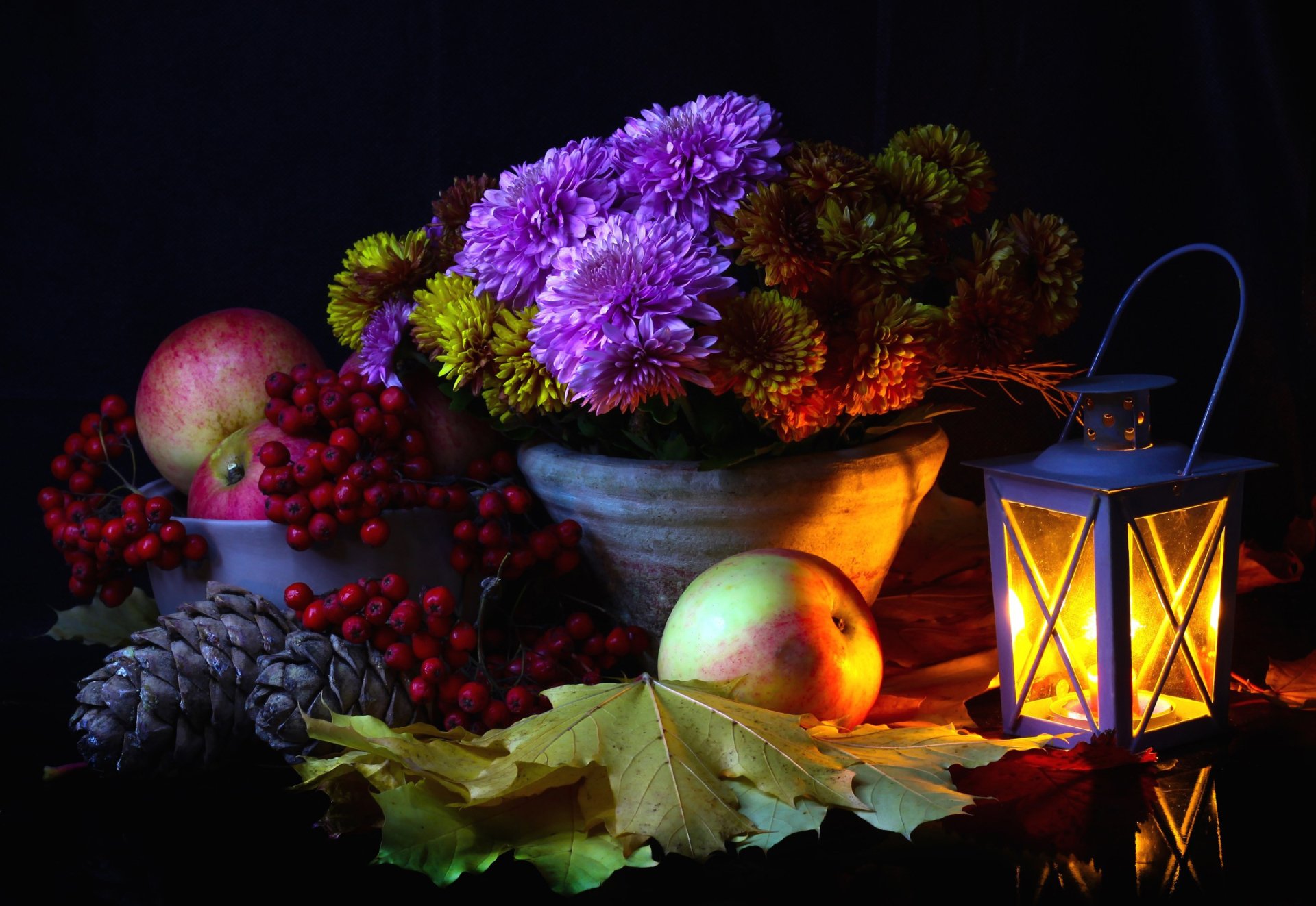 Still life HD wallpaper featuring a glowing lantern, bowl of chrysanthemums, apples, pine cones, berries, and autumn leaves in warm, low light photography.