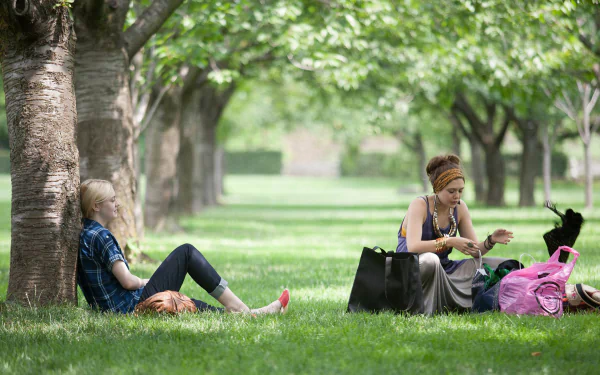 Elizabeth Olsen and Dakota Fanning sit under trees in a lush green park scene from the movie Very Good Girls, captured as an HD PC desktop wallpaper background.