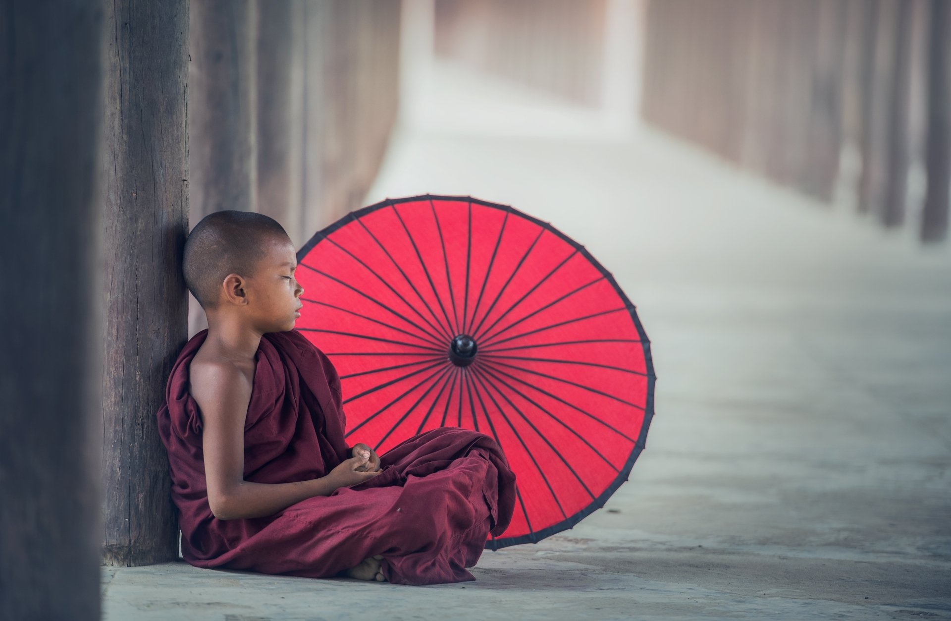 A serene young monk in robes sits beside a vibrant red umbrella, embodying tranquility in a Buddhist setting. This 4K Ultra HD wallpaper captures spiritual reflection and cultural beauty.