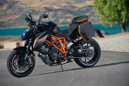 HD desktop wallpaper featuring a KTM 1290 Super Duke motorcycle with black and orange detailing parked on a scenic road near water and mountains.