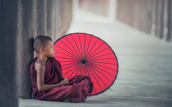 A serene young monk in robes sits beside a vibrant red umbrella, embodying tranquility in a Buddhist setting. This 4K Ultra HD wallpaper captures spiritual reflection and cultural beauty.