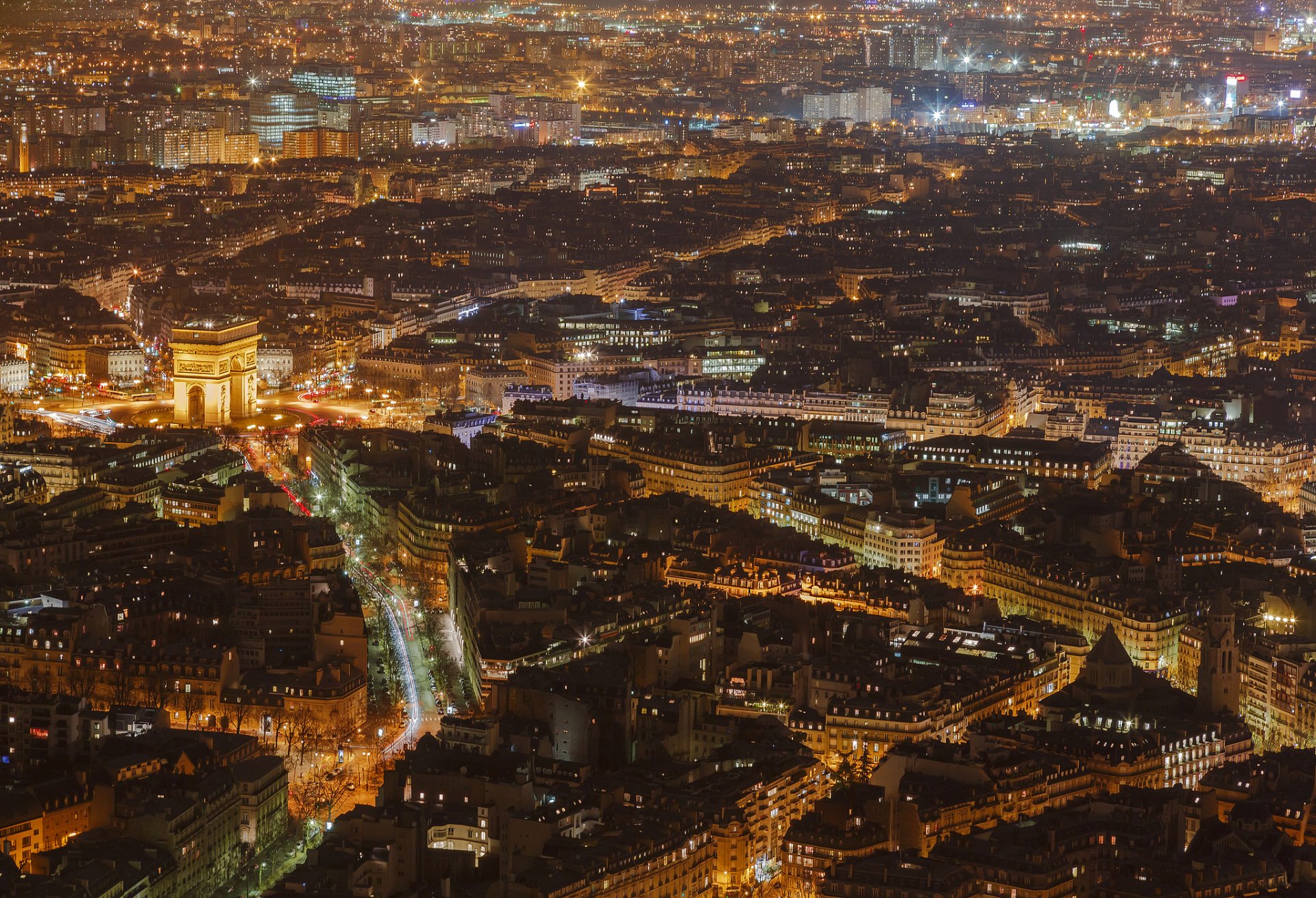 HD PC desktop wallpaper of Paris at night: aerial cityscape with the illuminated Arc de Triomphe, glowing city lights and dense man-made buildings.