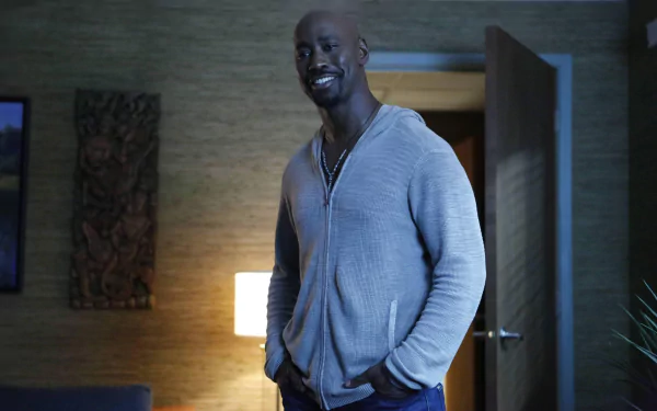 HD desktop wallpaper featuring Amenadiel from the TV show Lucifer, portrayed by D.B. Woodside, standing casually with a smile in a dimly lit room.