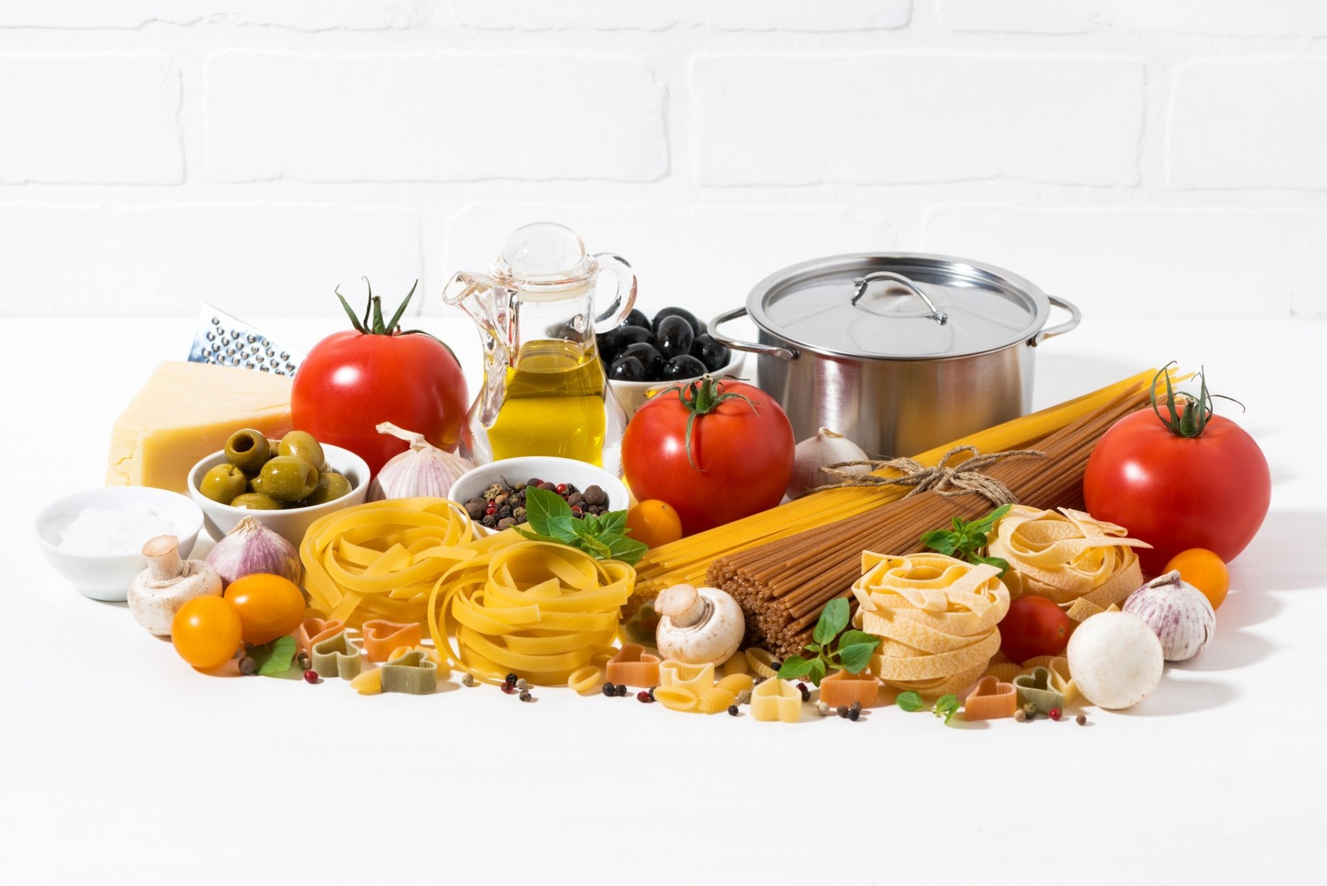 HD PC desktop wallpaper still life food scene: assorted dry spaghetti and nest pasta, tomatoes, cheese, olives, olive oil, mushrooms and a saucepan arranged on a white background.