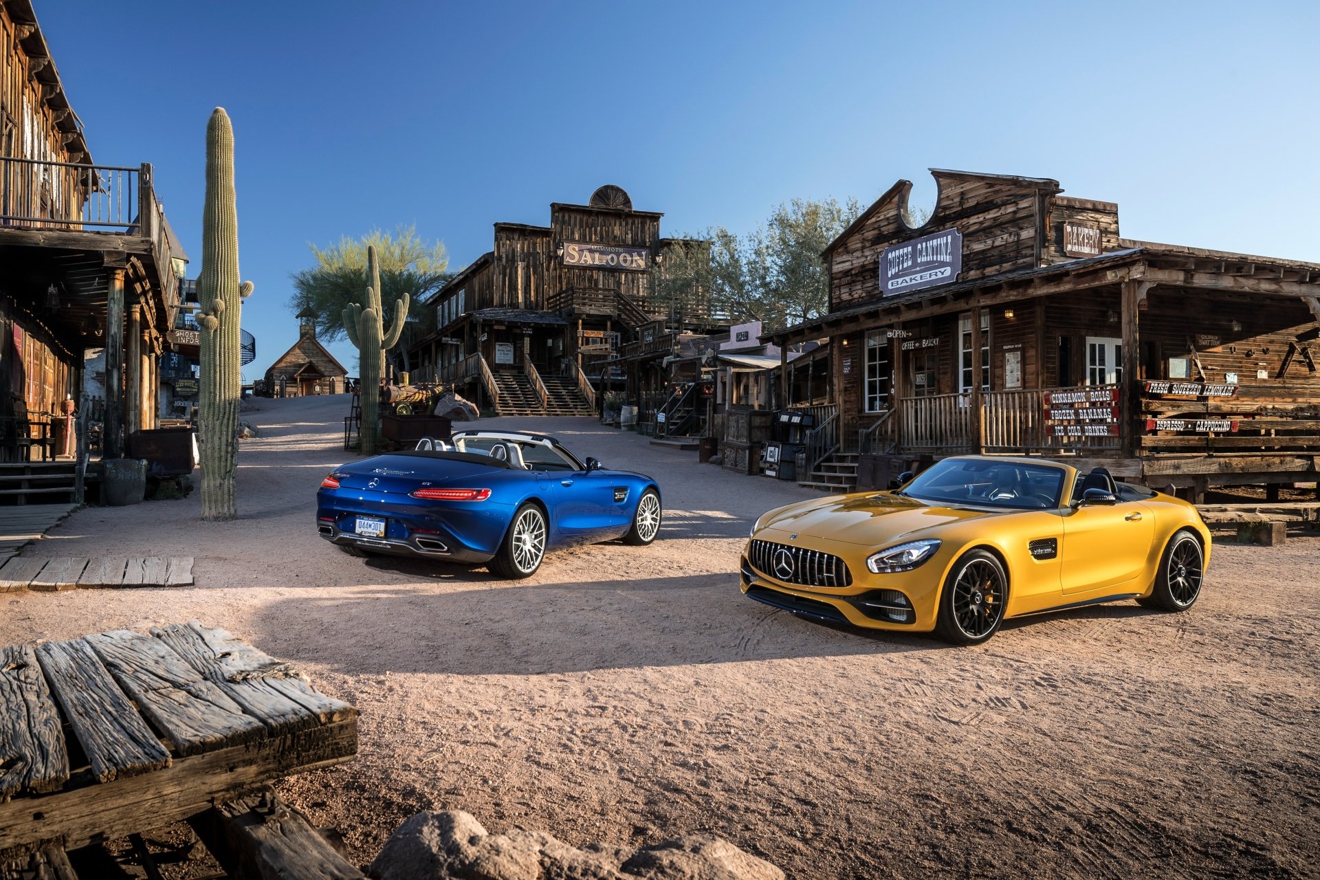Yellow Mercedes-AMG GT and blue convertible parked in a rustic old western town under clear blue sky, captured in 4K Ultra HD for PC desktop wallpaper.