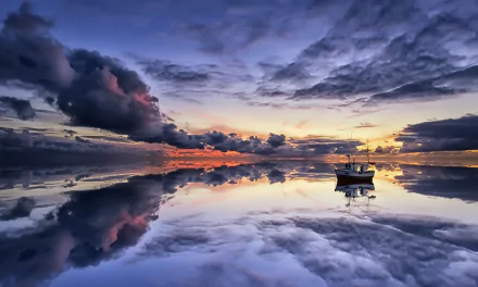 Fishing boat floats peacefully on a calm ocean at sunset, with dramatic clouds and sky perfectly reflected on the water, creating a serene horizon scene.