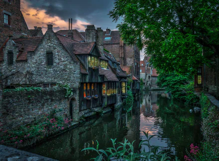 HD wallpaper of a charming canal in Brussels, Belgium. The scene features historic brick houses with flowers alongside the water. The dusk sky adds a serene ambiance to the man-made beauty.