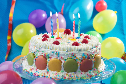 A festive HD desktop wallpaper featuring a colorful birthday cake adorned with macarons, whipped cream, raspberries, and candles. Vibrant balloons surround the cake, creating a celebratory atmosphere.