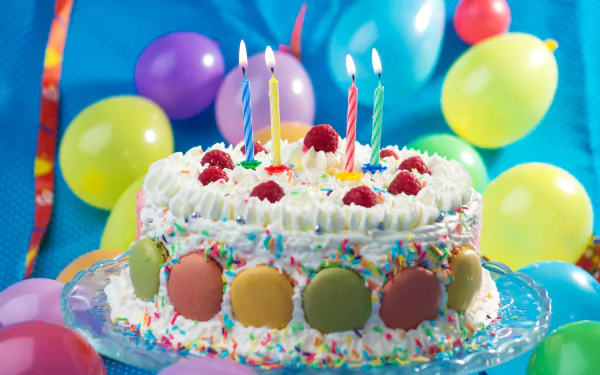 A festive HD desktop wallpaper featuring a colorful birthday cake adorned with macarons, whipped cream, raspberries, and candles. Vibrant balloons surround the cake, creating a celebratory atmosphere.