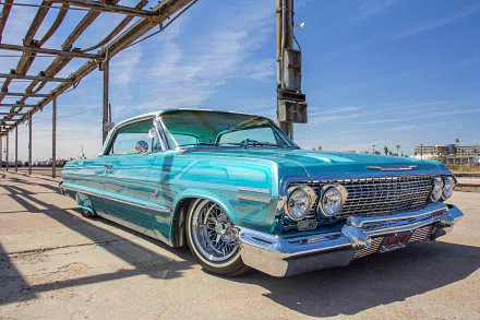 Turquoise classic Chevrolet Impala lowrider with chrome wire wheels and whitewall tires parked under industrial beams against a clear blue sky — HD desktop wallpaper.