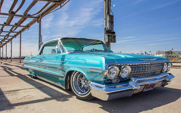 Turquoise classic Chevrolet Impala lowrider with chrome wire wheels and whitewall tires parked under industrial beams against a clear blue sky — HD desktop wallpaper.