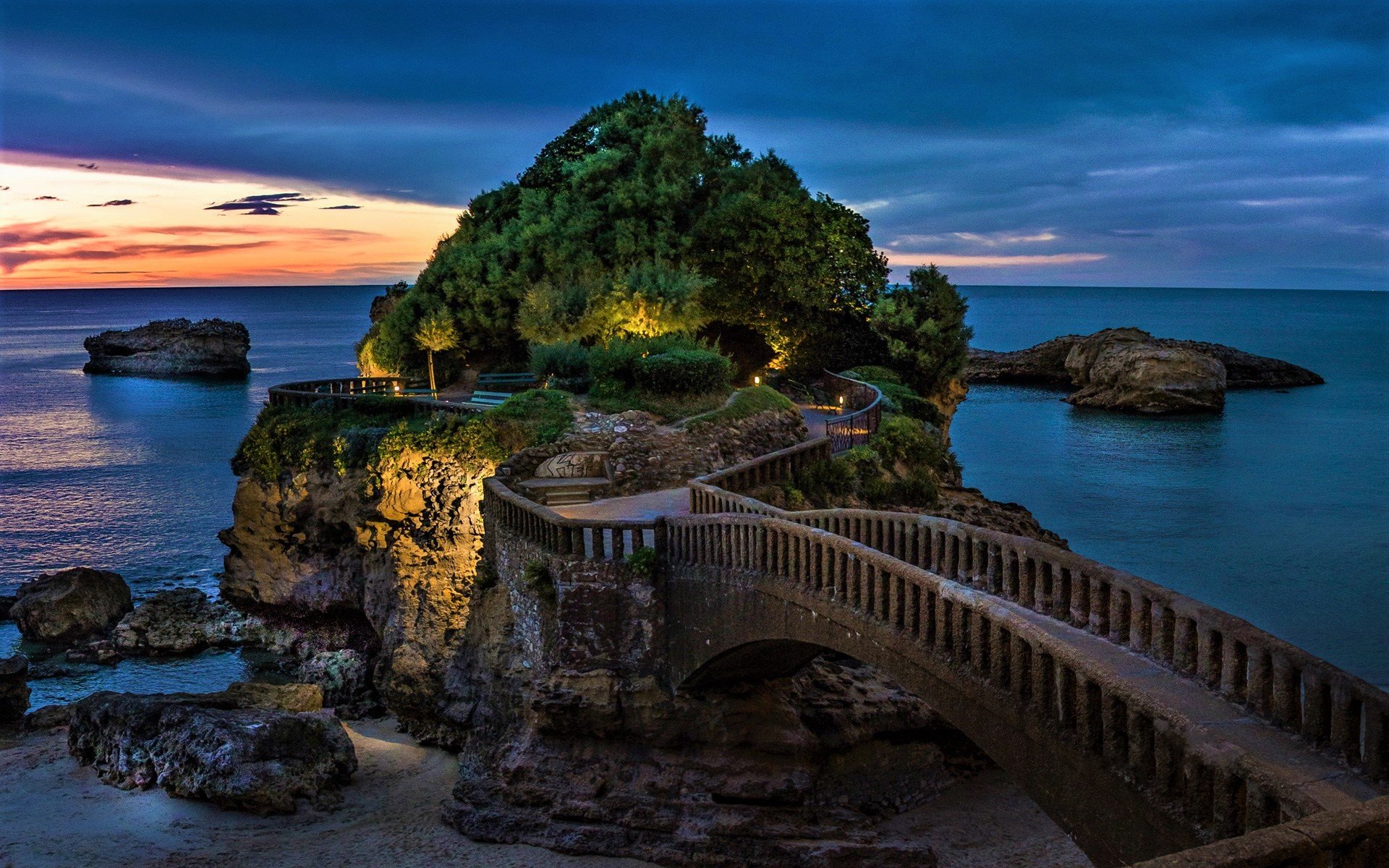 HD desktop wallpaper of Roca de Basta, France, featuring a beautifully lit man-made bridge extending towards a lush, green island against a serene ocean at sunset.