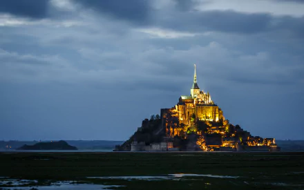 Illuminated Mont Saint-Michel abbey at dusk rising from tidal flats, a religious landmark captured as an HD PC desktop wallpaper and background.