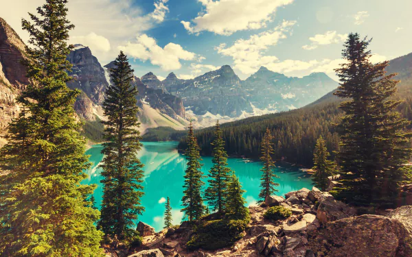 Moraine Lake in Banff National Park, Canada, showcases turquoise water surrounded by evergreen trees, forest, and rugged mountain peaks under a partly cloudy sky.