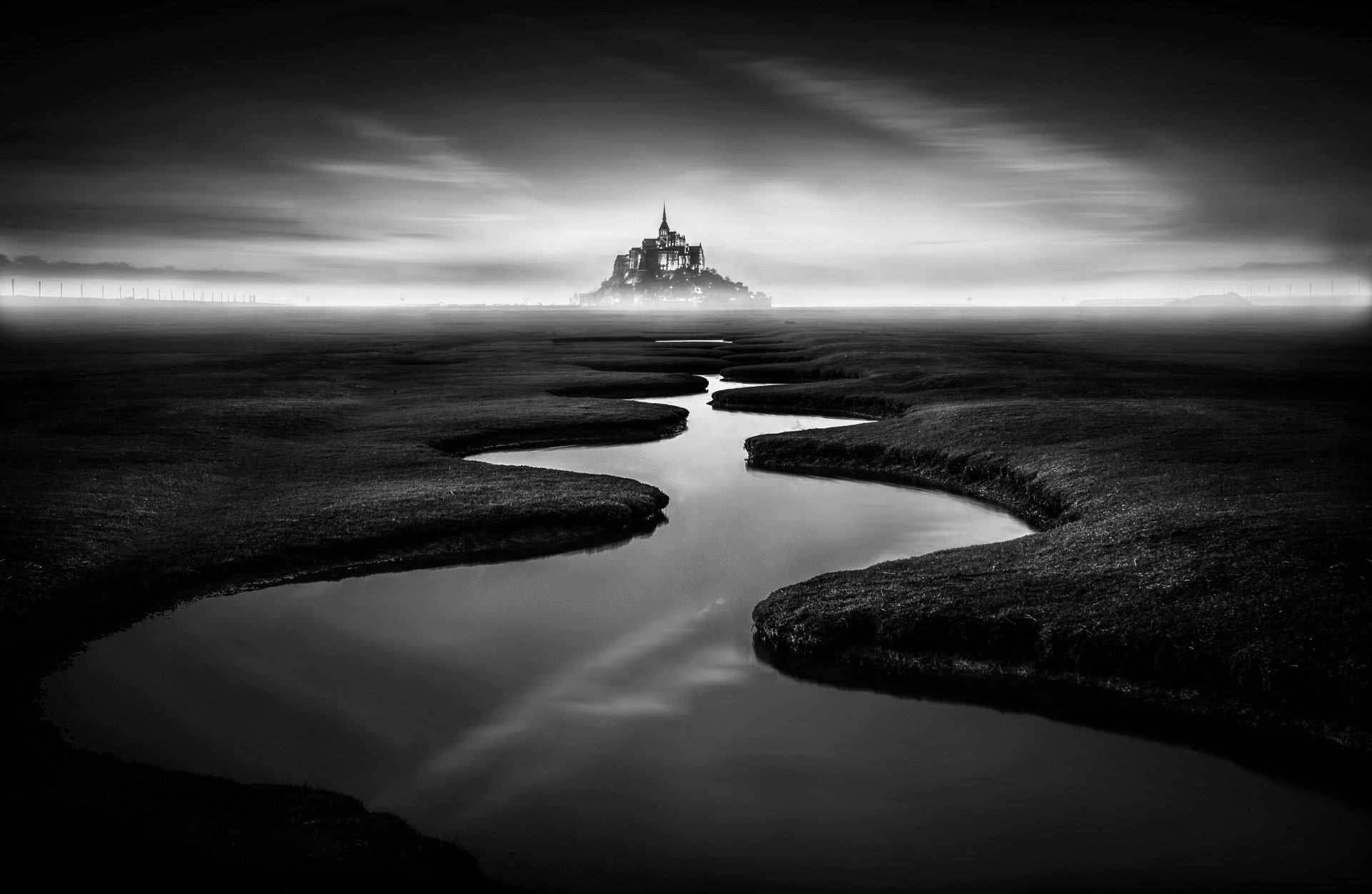 Black and white 4K landscape of Mont Saint-Michel in France, shrouded in fog with a reflective waterway leading to the religious landmark.