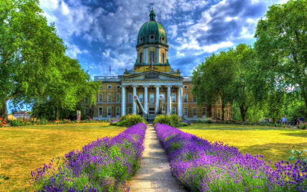 Vivid HDR 4K image of the Imperial War Museum in spring, framed by green trees and a vibrant purple flower garden along a central path.