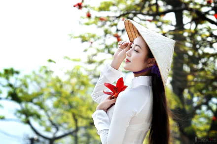 A Vietnamese woman wearing a traditional Ao Dai and an Asian conical hat stands amidst blooming flowers with a soft bokeh background, captured in HD for a desktop wallpaper.