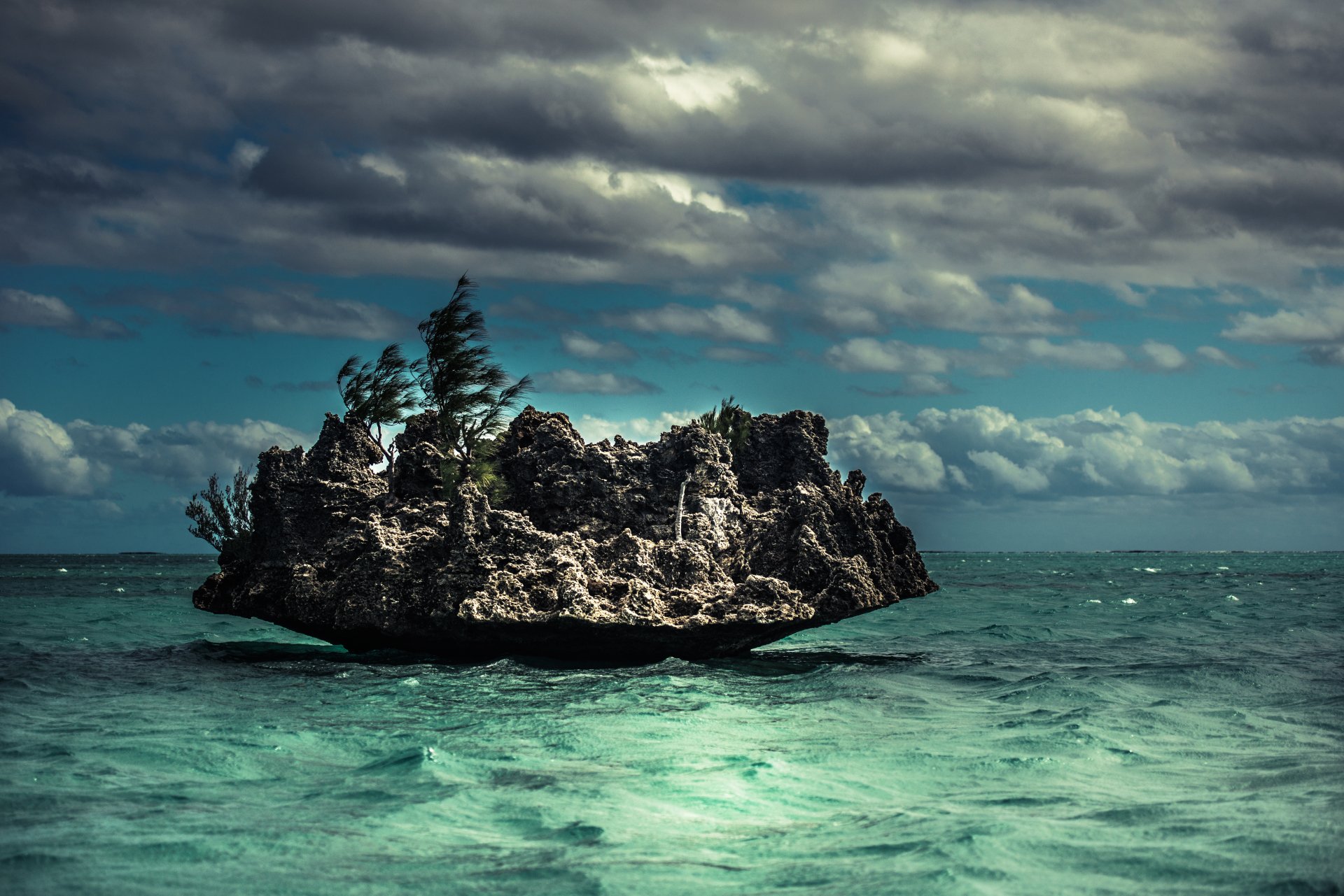 HD desktop wallpaper featuring a seascape with waves crashing against a rocky outcrop. The ocean is vibrant under a cloudy sky with wind-blown trees on the rock and calm sea around.