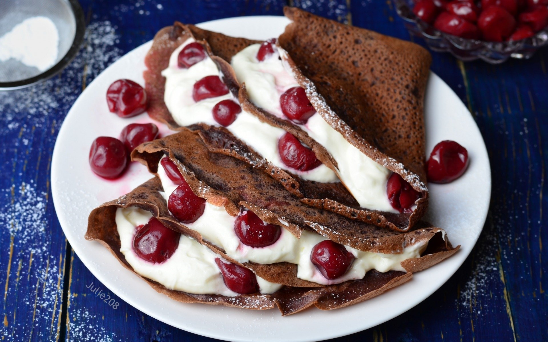 HD desktop wallpaper showing chocolate crêpes filled with cream and topped with fresh cherries on a white plate against a blue wooden surface.