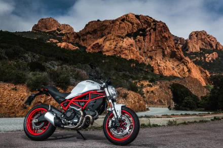 A Ducati Monster motorcycle with red frame and white body parked on a road against a backdrop of rugged, sunlit rocky mountains in 4K Ultra HD detail.