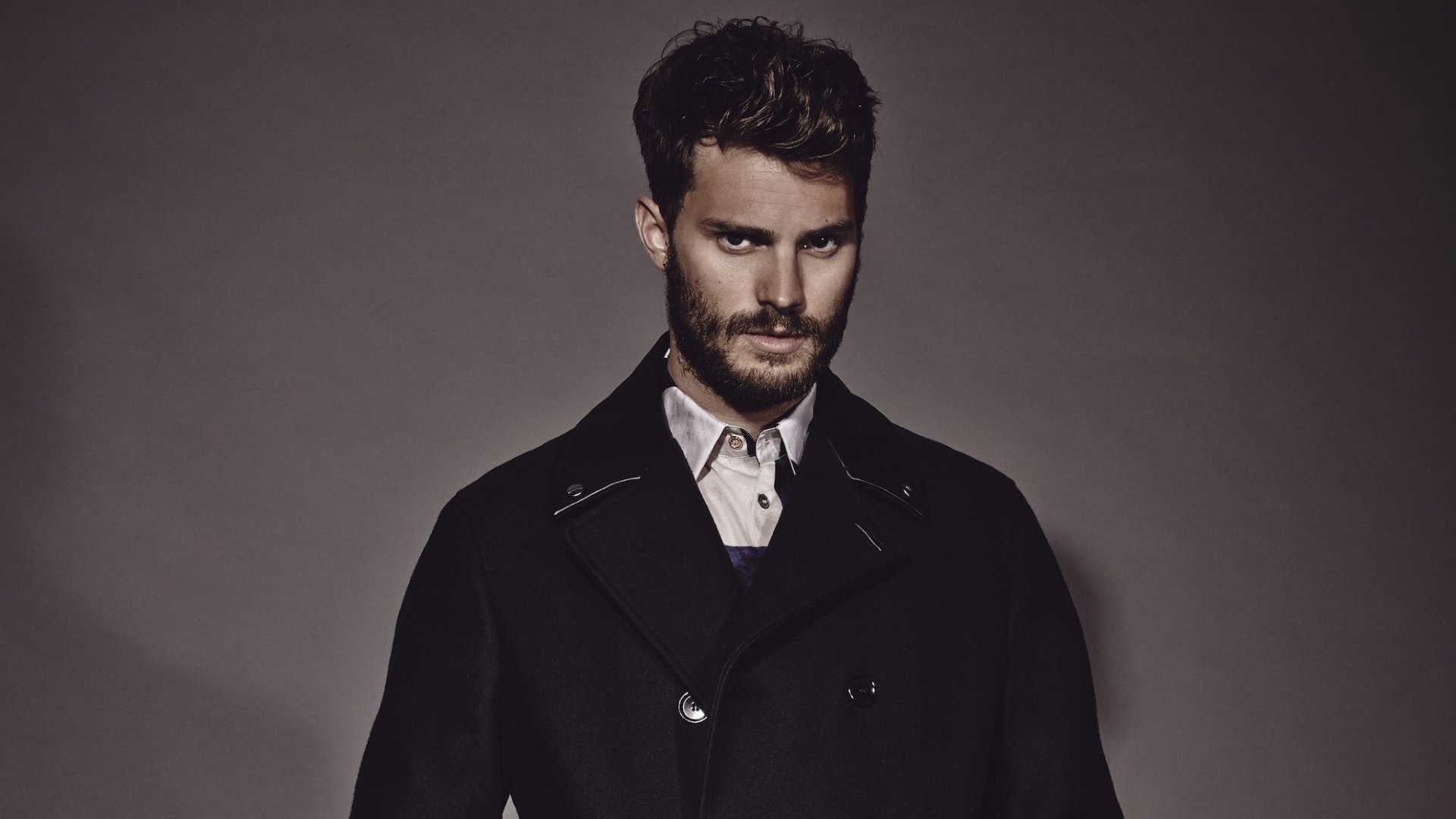 A striking HD desktop wallpaper featuring celebrity Jamie Dornan, showcasing his stylish look with a beard and a dark coat against a neutral background.