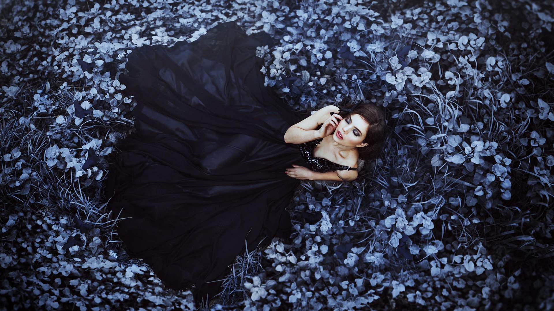 A brunette woman in a flowing black dress lies amidst blue-toned foliage, exuding a serene and moody atmosphere in this HD desktop wallpaper and background.