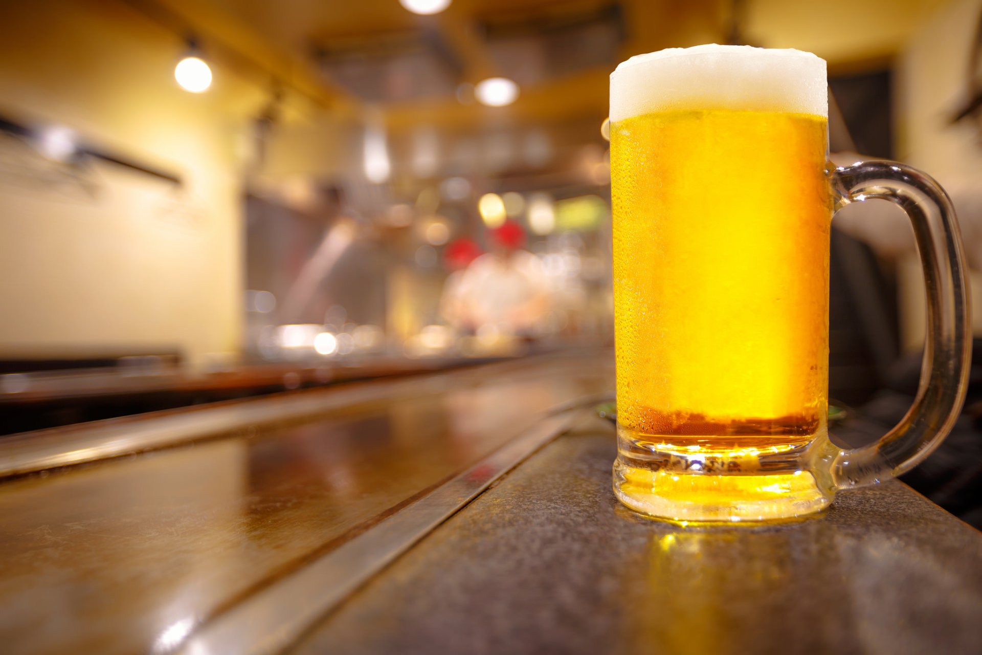 A frosty glass of beer sits on a bar counter with a blurred background, showcasing depth of field. This HD wallpaper highlights the golden hue and foamy head, embodying a refreshing alcoholic beverage.