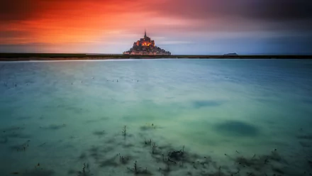 Mont Saint-Michel monastery in France glows at sunset, reflected in the calm ocean waters, captured in stunning HD detail as a desktop wallpaper background.