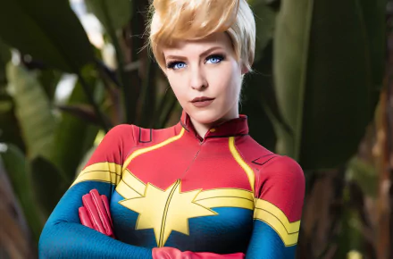 A woman with blue eyes in a Captain Marvel cosplay outfit stands confidently. This HD desktop wallpaper and background showcases detailed costume design and vibrant colors.