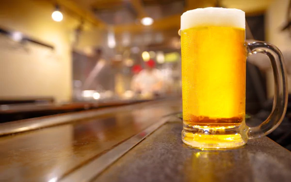 A frosty glass of beer sits on a bar counter with a blurred background, showcasing depth of field. This HD wallpaper highlights the golden hue and foamy head, embodying a refreshing alcoholic beverage.