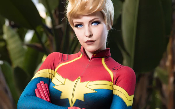A woman with blue eyes in a Captain Marvel cosplay outfit stands confidently. This HD desktop wallpaper and background showcases detailed costume design and vibrant colors.