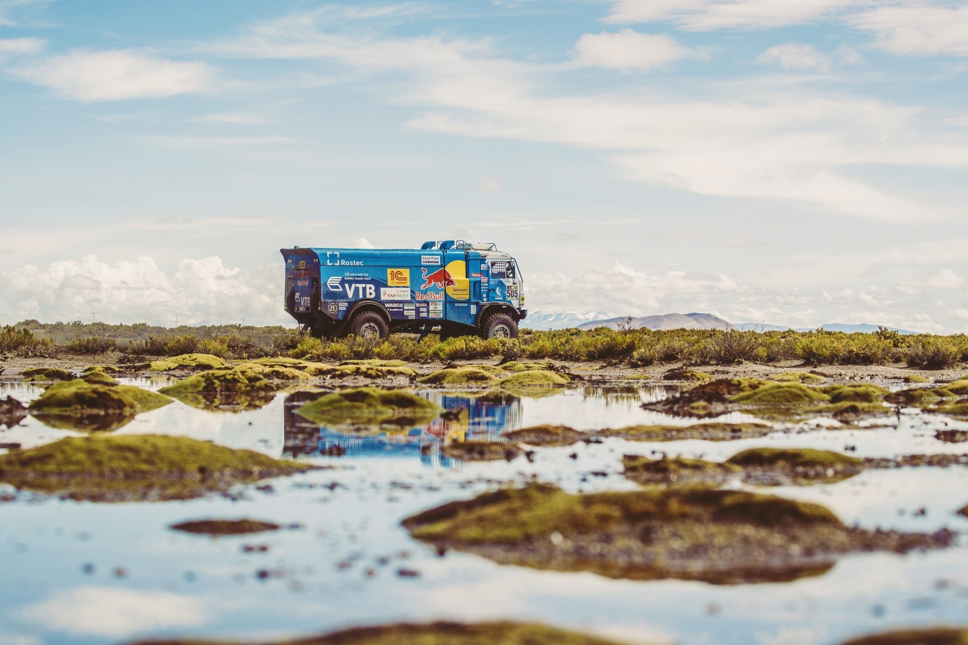 A Kamaz rally truck with Red Bull branding drives across a rocky landscape under a blue sky, its reflection visible in the water below, capturing dynamic sports action.