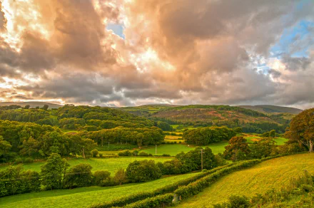 HD desktop wallpaper of a lush Welsh landscape, featuring vibrant green fields, dense forests, and dramatic clouds under a colorful sky.