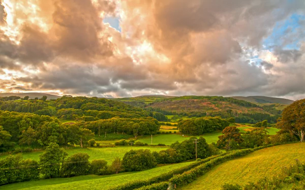 HD desktop wallpaper of a lush Welsh landscape, featuring vibrant green fields, dense forests, and dramatic clouds under a colorful sky.