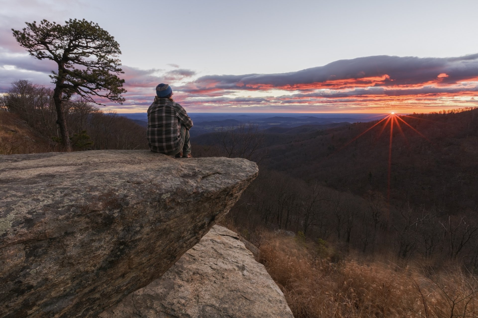 Sunrise over Shenandoah National Park in Virginia, USA, with a person sitting on a rock ledge overlooking the expansive morning landscape.