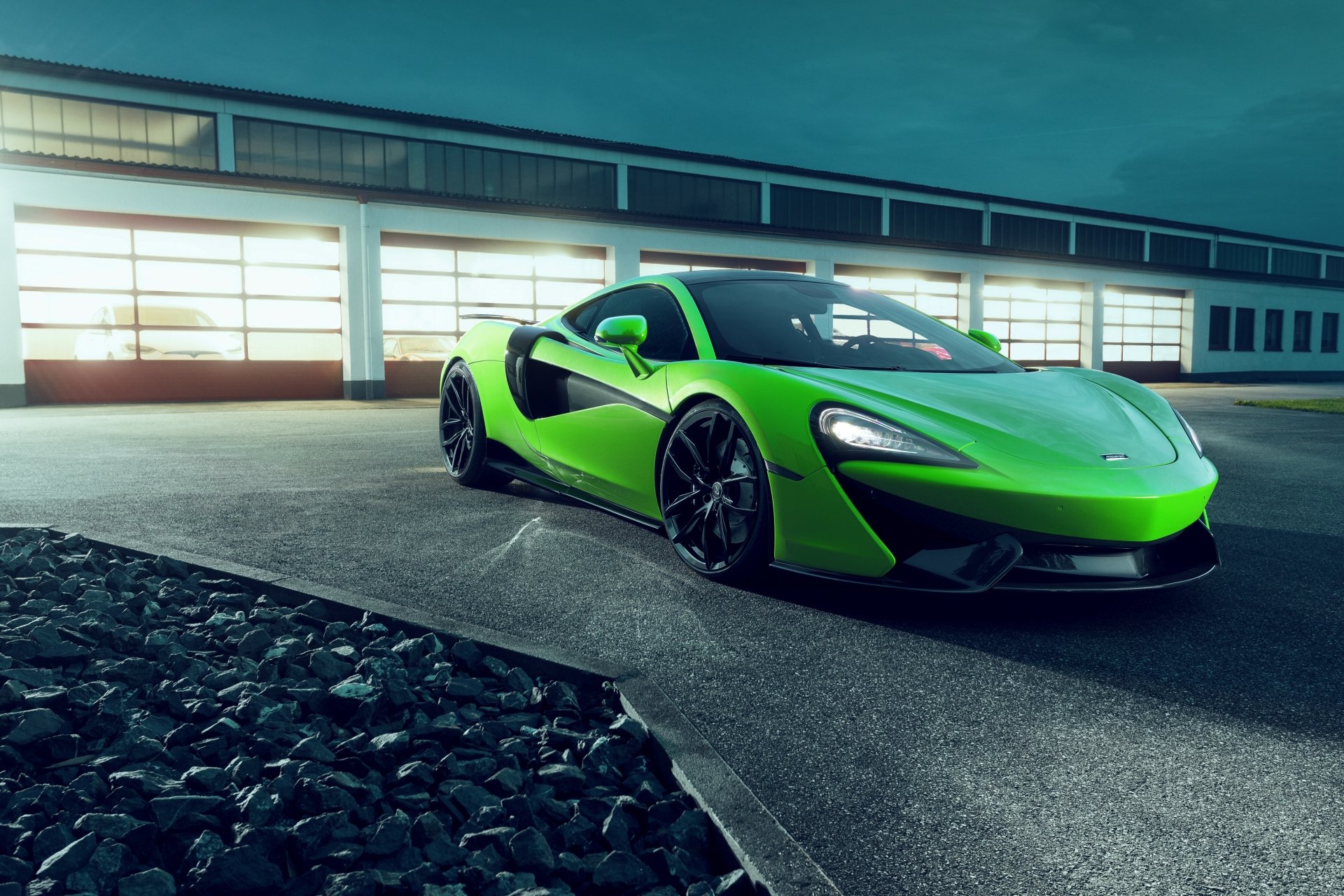 A vibrant green McLaren 570GT supercar coupe showcased in a 4K Ultra HD desktop wallpaper, parked in front of a modern industrial building at dusk.