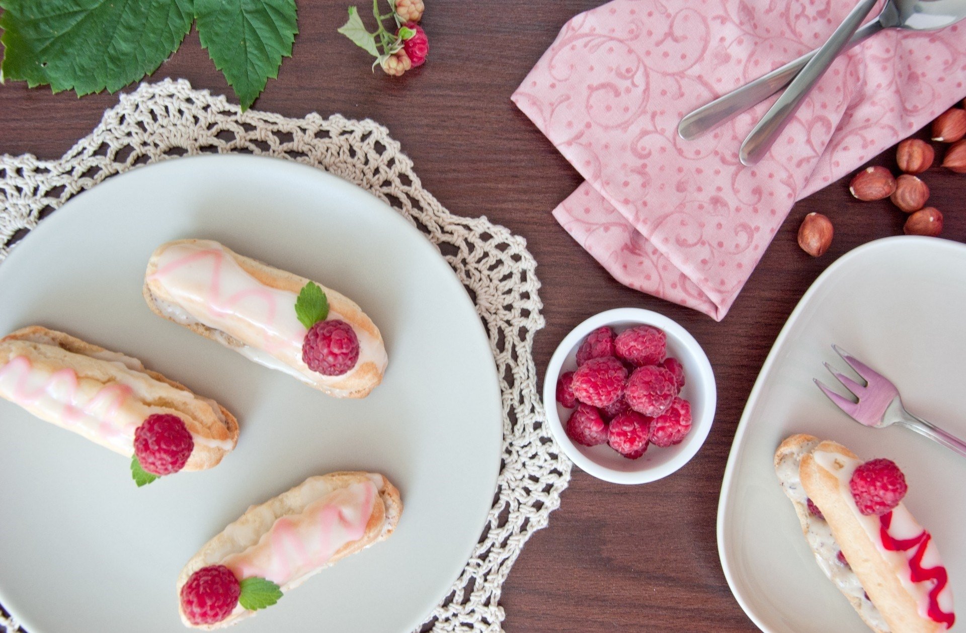 HD PC desktop wallpaper background — still life of raspberry éclairs on white plates with doily, bowl of raspberries, pink napkin and scattered hazelnuts; pastry dessert close-up.