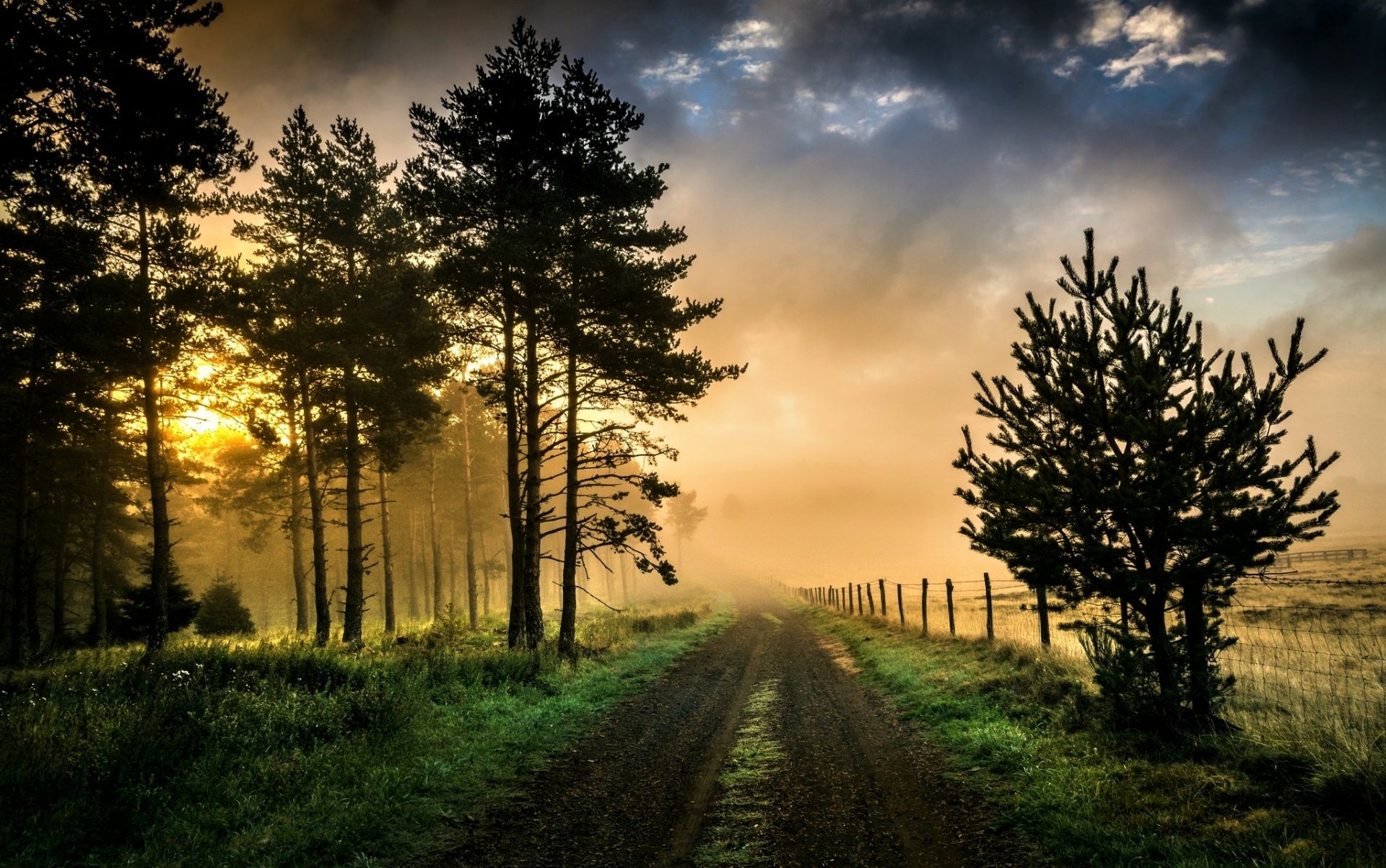 A foggy country dirt road flanked by a fence and trees, stretching through a grassy field under a dramatic sky, creating a serene HD desktop wallpaper scene.