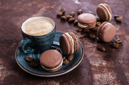 A HD desktop wallpaper featuring a still life of coffee beans, a cup of frothy coffee on a blue plate, and several chocolate macarons.
